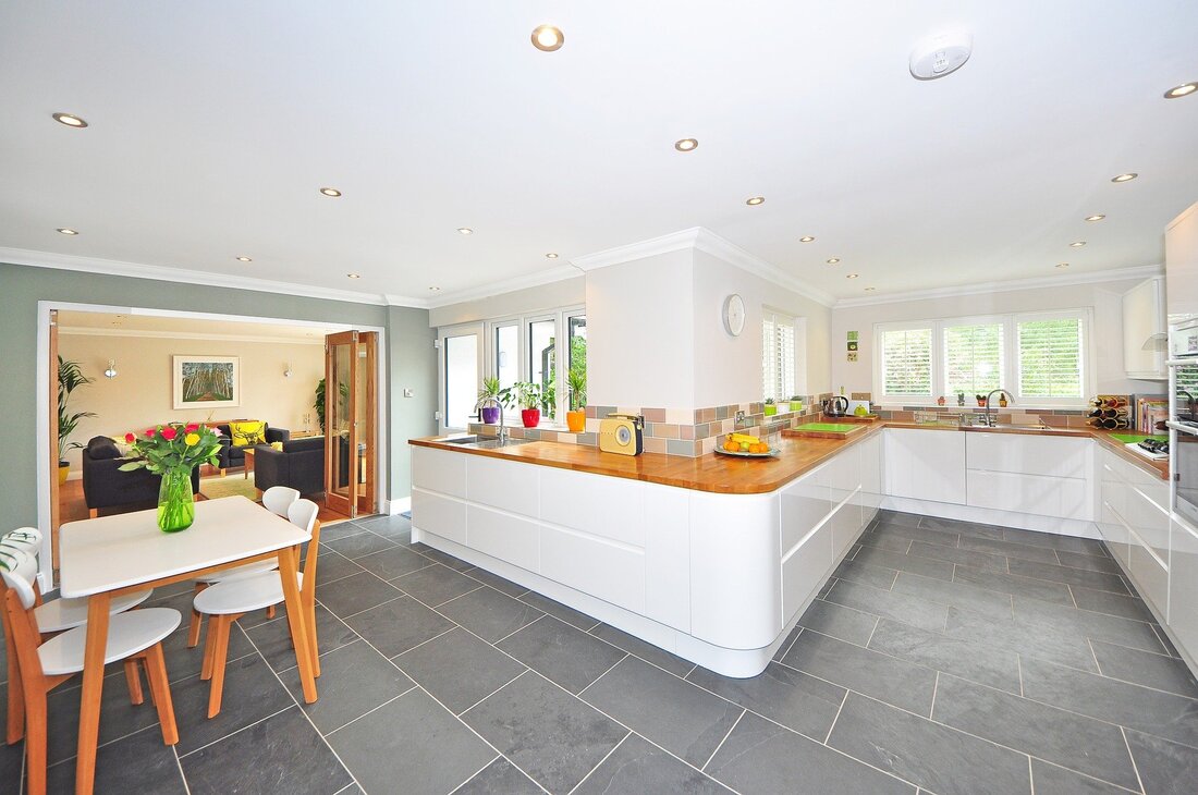 Large open plan kitchen diner, with white kitchen units, grey floor tiles, and timber worktops. The walls are painted white and the rear wall is light green