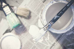 Birds eye picture of a paint tin filled with white paint. There is a paint brush resting on top of the tin and other paint brushes lying on on some newspaper