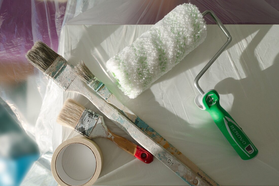 Picture of a paint roller with different sized brushes and masking tape laid on dust sheets.