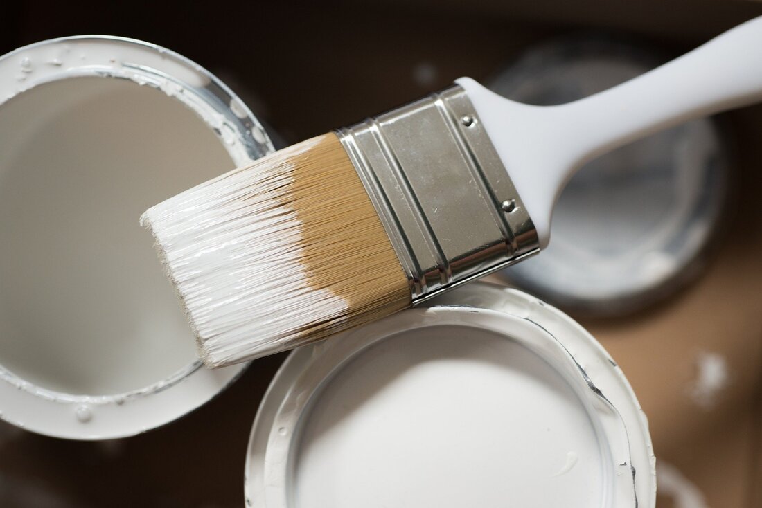 Birds eye view picture of two paint tins filled with white paint with a 2 inch paint brush resting on top